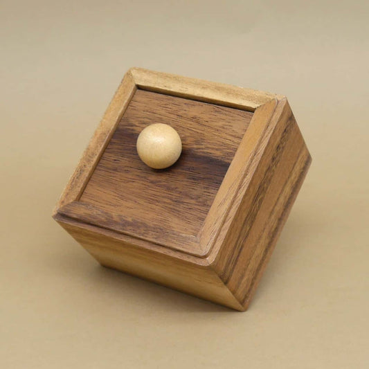 Quadratische Geschenkbox aus gemasertem Holz mit hellem Kugelknauf, leicht gekippt vor sandfarbenem Hintergrund.